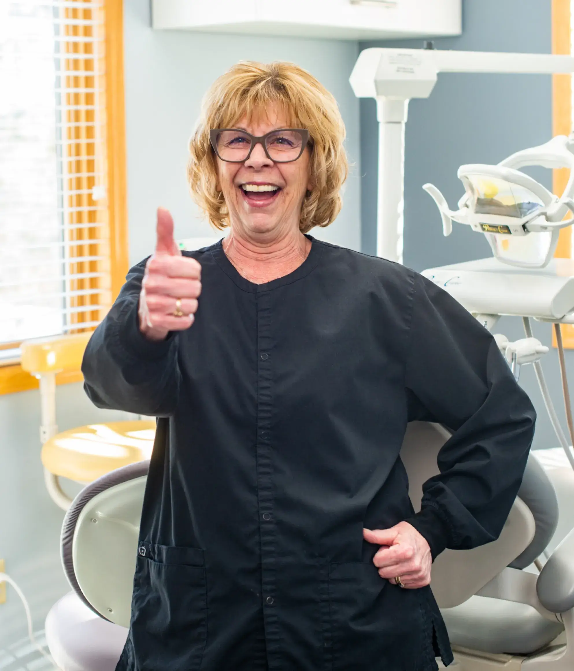 Smiling woman in a dental office giving a thumbs-up while wearing a black coat.